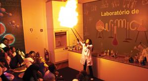 Descrição da imagem: laboratório de química. A esquerda, crianças sentadas de perfil observando uma mulher à direita, de óculos de proteção, jaleco branco e calças preta, as 2 mãos estão esticadas para o alto segurando um bastão em cada mão, e neles está saindo fogo.