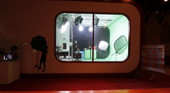 Descrição da imagem. Através de um vidro retangular se observa um estúdio de televisão, com luzes, projetores e um homem sentado à frente de um computador.