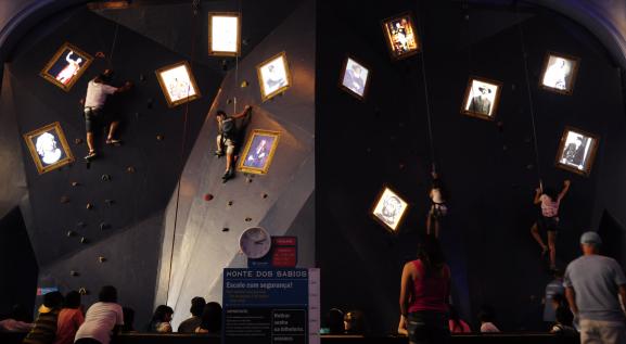 Descrição da imagem: Quatro pessoas escalando em uma parede e contém quadros de artistas. Pessoas de costas os observam, no centro uma placa azul, escrito em letra branca: Monte dos sábios Escale com segurança.