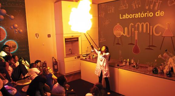 Descrição da imagem: laboratório de química. A esquerda, crianças sentadas de perfil observando uma mulher à direita, de óculos de proteção, jaleco branco e calças preta, as 2 mãos estão esticadas para o alto segurando um bastão em cada mão, e neles está saindo fogo.