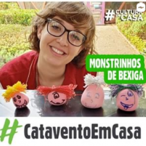 Foto de uma mulher com jaleco vermelho e óculos sorrindo. Na sua frente uma mesa com alguns objetos na cor rosa. Ao fundo o borboletário do Museu Catavento com algumas plantas