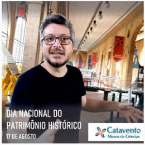 Homem com camiseta preta e óculos sorrindo e gesticulando com as mãos. Ao fundo seção engenho do Museu Catavento