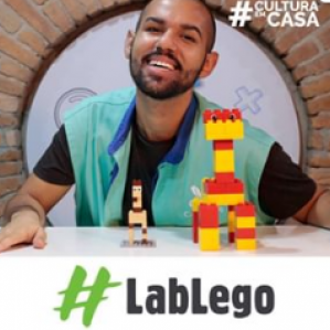Homem de colete azul sorrindo. Na sua frente uma mesa branca com algumas peças de lego em cima