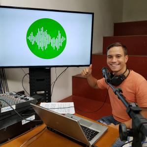 Foto de homem jovem, branco, em frente a um notebook e equipamentos de rádio. Ao fundo uma tela com o logotipo do programa "Frequências da Ciência".