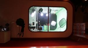 Descrição da imagem. Através de um vidro retangular se observa um estúdio de televisão, com luzes, projetores e um homem sentado à frente de um computador.